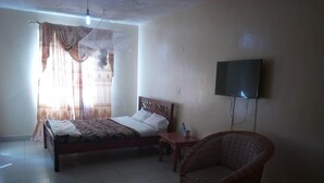 A bedroom with a bed, a chair, a small table, a TV, and a window with curtains. - Bellview Hotel (Thika)