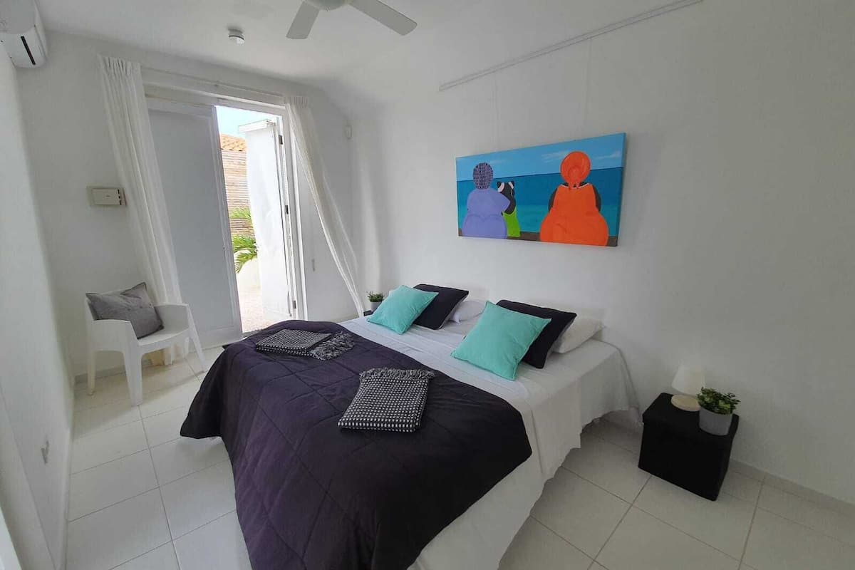 A bedroom with a bed, a ceiling fan, a painting, a chair, and a small table with a plant.