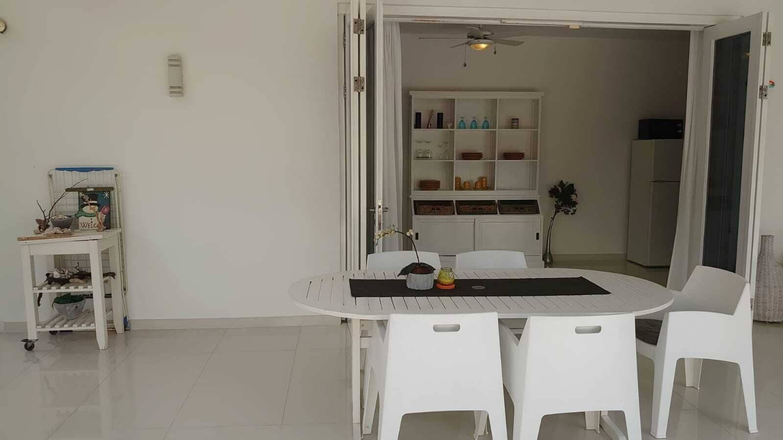 A dining area with a white table and chairs, a small side table with a 'Welcome' sign, and a white cabinet with shelves in the background.