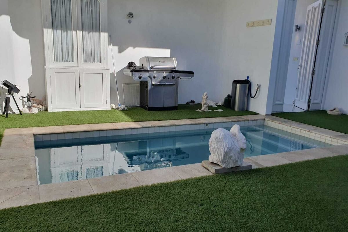 A swimming pool with a stone border, a grill, and a statue in a backyard.
