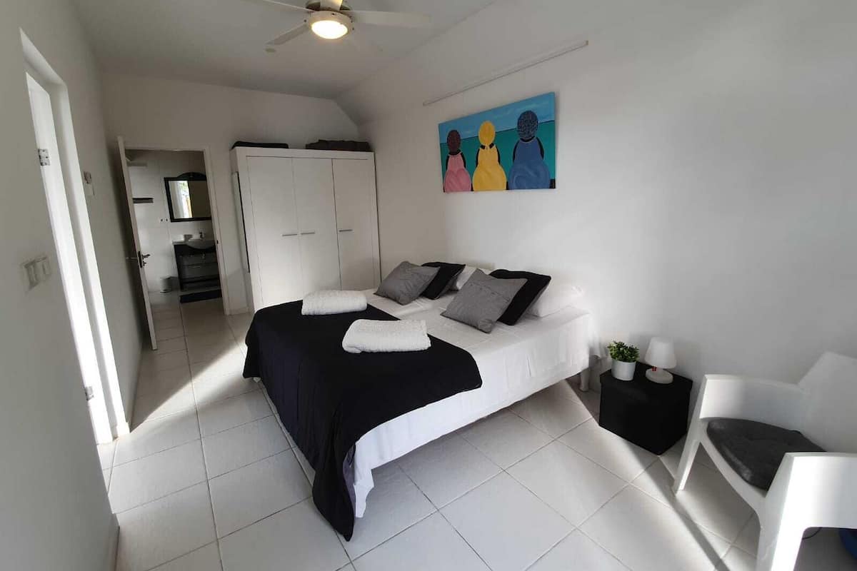 A bedroom with a bed, a ceiling fan, a painting, and a small table with a lamp.