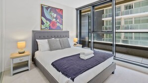 A modern bedroom with a large bed, a nightstand with a lamp, a decorative wall piece, and a large window with a view of other buildings. - YARRA · YARRA Toorak Rd & Chapel St. Pool, Gym & Parking (South Yarra)