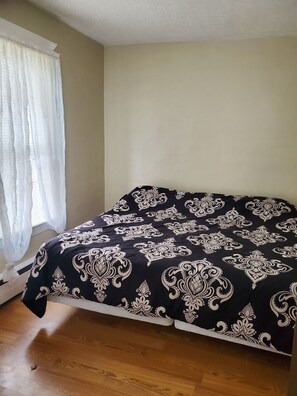 Une chambre à coucher avec un lit recouvert d’une couvre-lit à motifs noir et blanc, un plancher en bois et une fenêtre ornée de rideaux légers.