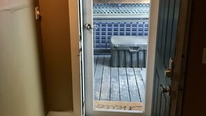 A narrow hallway with a wooden floor leading to a door. The door opens to a patio area with a table and chairs, a wooden fence, and a view of a building with a tiled roof.