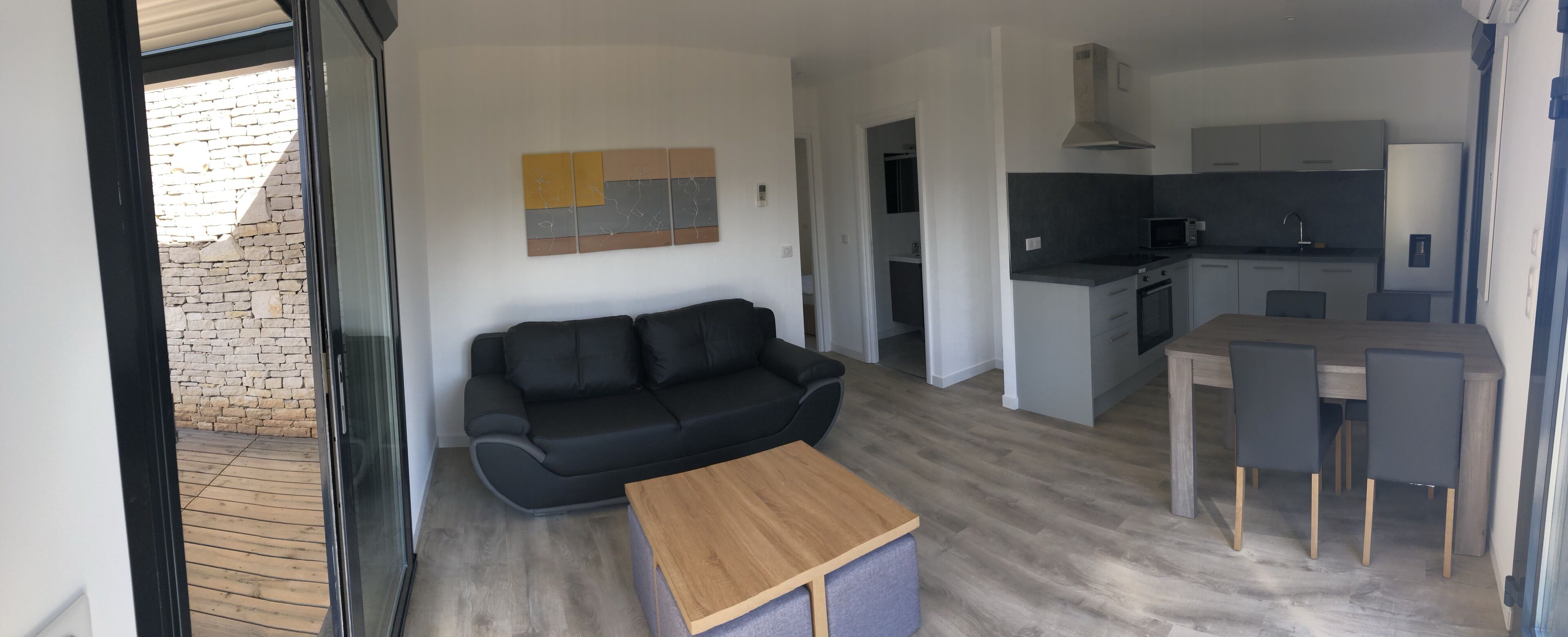 Una sala de estar moderna con un sofá negro, una mesa de centro de madera y un sillón de acento gris. Se ve una cocina con gabinetes grises y un microondas al fondo.