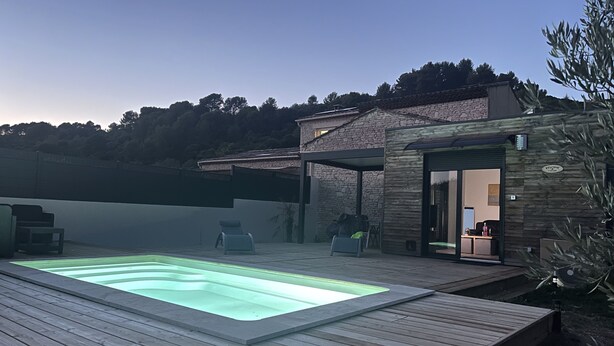 Eine Holzterrasse mit Schwimmbecken, ein Steinhaus mit Holzfassade und Blick auf Bäume im Hintergrund.