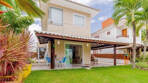 A two-story house with a covered patio, a lawn, and a small garden area. - GB07 Otima Casa 4 Suites em Guarajuba (Camaçari)