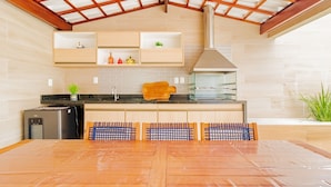 A modern kitchen with wooden cabinets, a built-in oven, and a large dining table with chairs. - GB07 Otima Casa 4 Suites em Guarajuba (Camaçari)