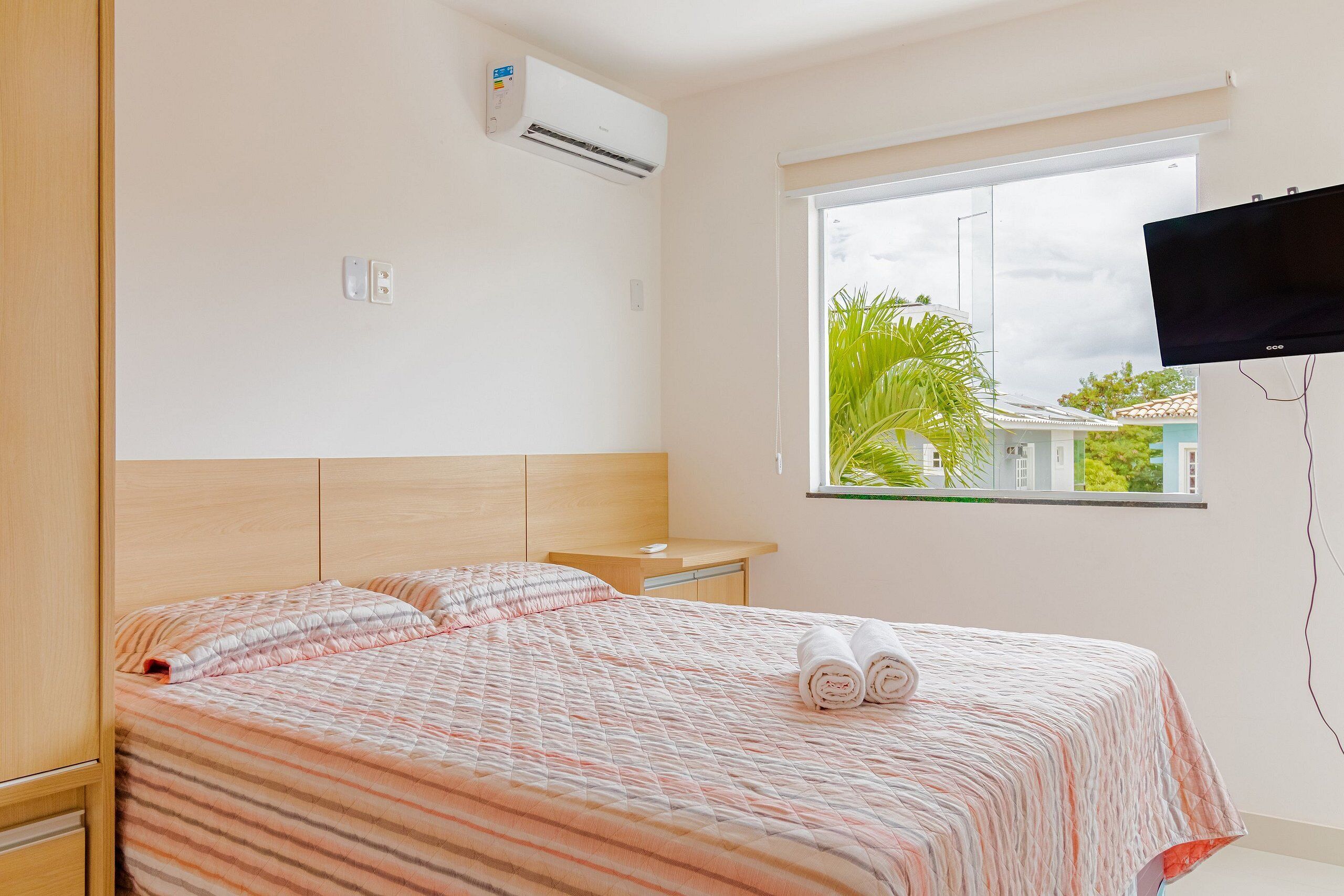 A hotel room with a bed, a TV mounted on the wall, and a window with a view of greenery outside.
