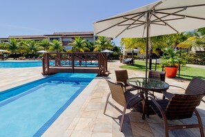 A pool area with a lounge chair and umbrella, surrounded by palm trees and a building in the background. - GB11 Otimo Ap Genipabu Sum Residence (Camaçari)