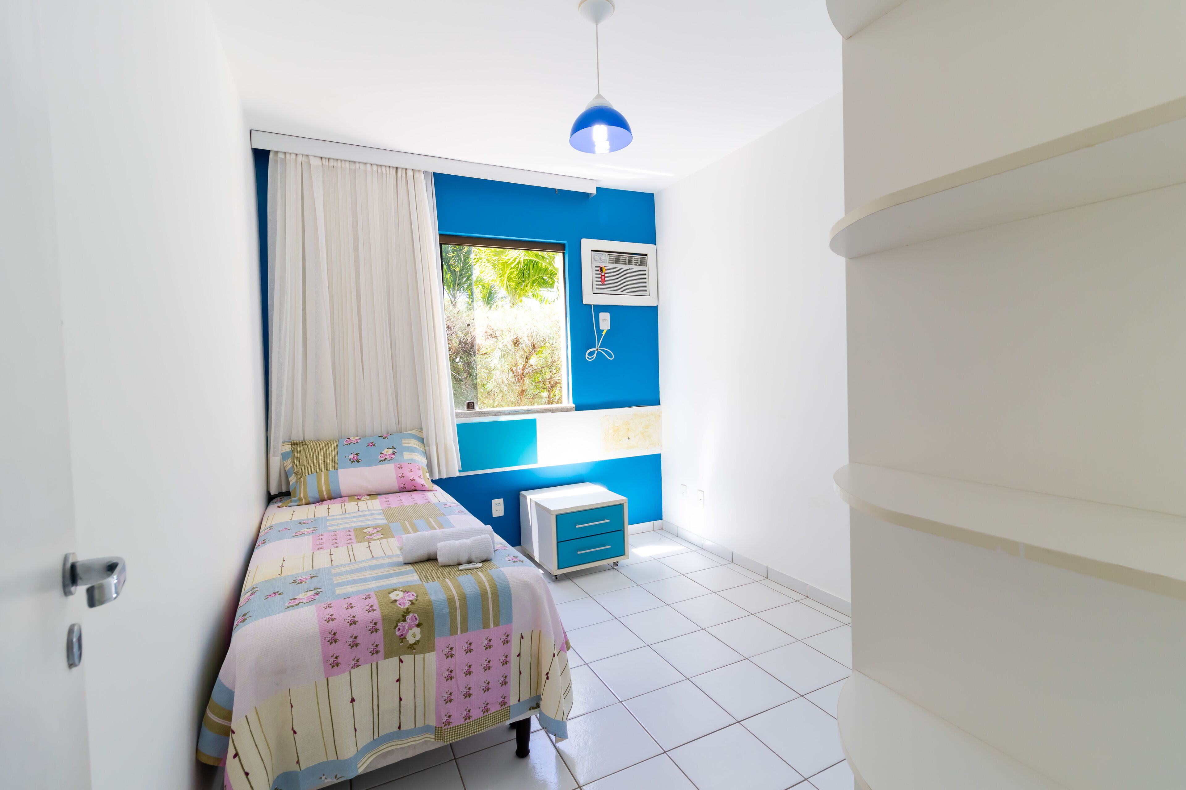 Ein Schlafzimmer mit Bett, Nachttisch, einem Fenster mit Vorhängen, Deckenleuchte und Wand-Klimaanlage.