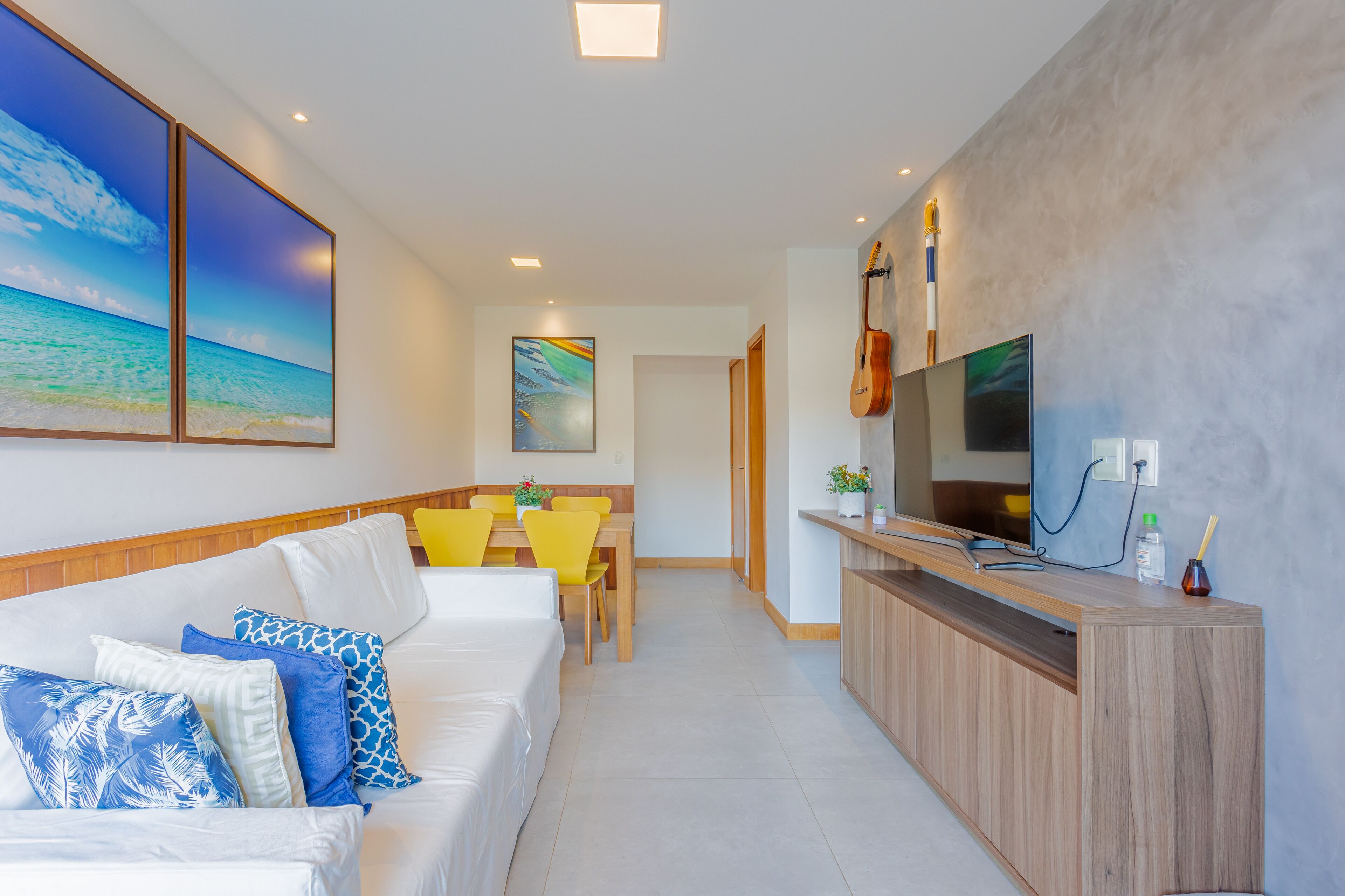 Quarto de hotel moderno com sofá branco, cadeiras amarelas, aparador de madeira e pintura com paisagem de praia.
