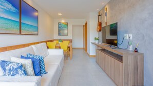 A modern hotel room with a white sofa, yellow chairs, a wooden TV stand, and a painting of a beach scene.