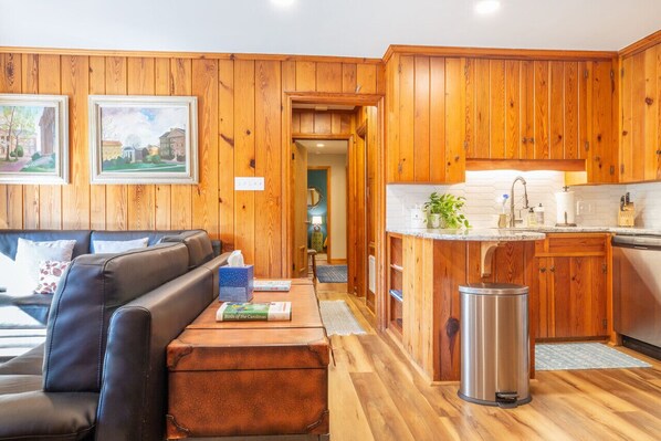 A cozy living room with a leather sofa, wooden walls, and a kitchen area with wooden cabinets and a stainless steel trash can.