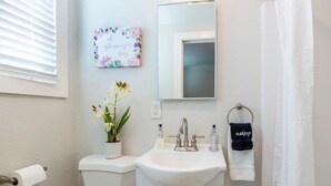 A bathroom with a toilet, sink, and mirror. There is a floral sign on the wall and a towel rack with a towel. - @ Marbella Lane - MiniGuest House | DTWN SJ | Parking (San Jose)