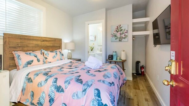 A bedroom with a wooden headboard, a bed with a pink and blue leaf-patterned bedspread, a white bedside table with a lamp, and a red door. - @ Marbella Lane - MiniGuest House | DTWN SJ | Parking (San Jose)