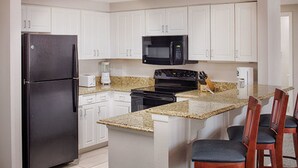 A kitchen with white cabinets, a black refrigerator, a microwave, a stove, and a granite countertop. There are two wooden chairs with cushioned seats. - Two Bedroom, Lock Off, Ocean Front, Luxury Condo, Daytona Beach, FL 1948956 (Daytona Beach)