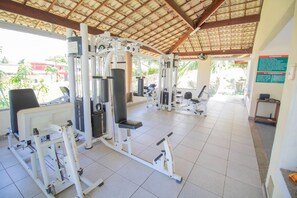 A gym with various exercise machines, a ceiling fan, and a view of the outdoors. - VT01 Casa 4 Suites em Vilas do Atlantico (Lauro de Freitas)