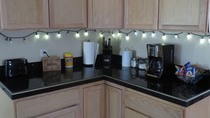 Cozinha com armários de madeira, bancada preta, cafeteira, torradeira e luzes de cordão.