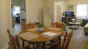 Uma sala de jantar com mesa de madeira, quatro cadeiras, um espelho grande e uma televisão ao fundo.