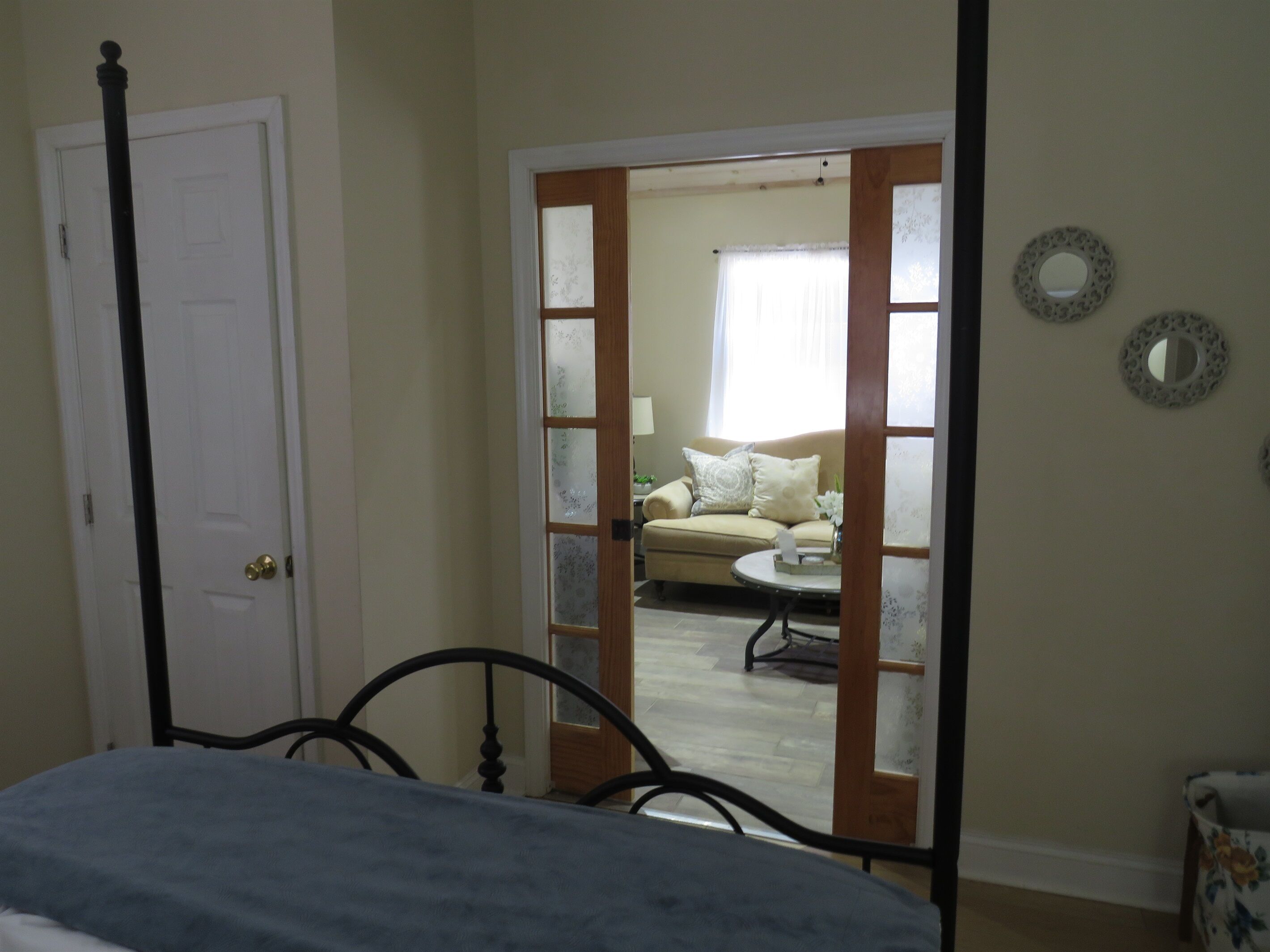 A bedroom with a four-poster bed, a door, and a view into a living room with a sofa and a coffee table.