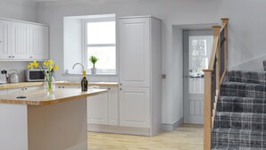 Una cucina moderna con mobili bianchi, un piano di lavoro in legno e una piccola isola. Si vede una finestra con vista sull'esterno e una scala rivestita con un tappeto a quadri.
