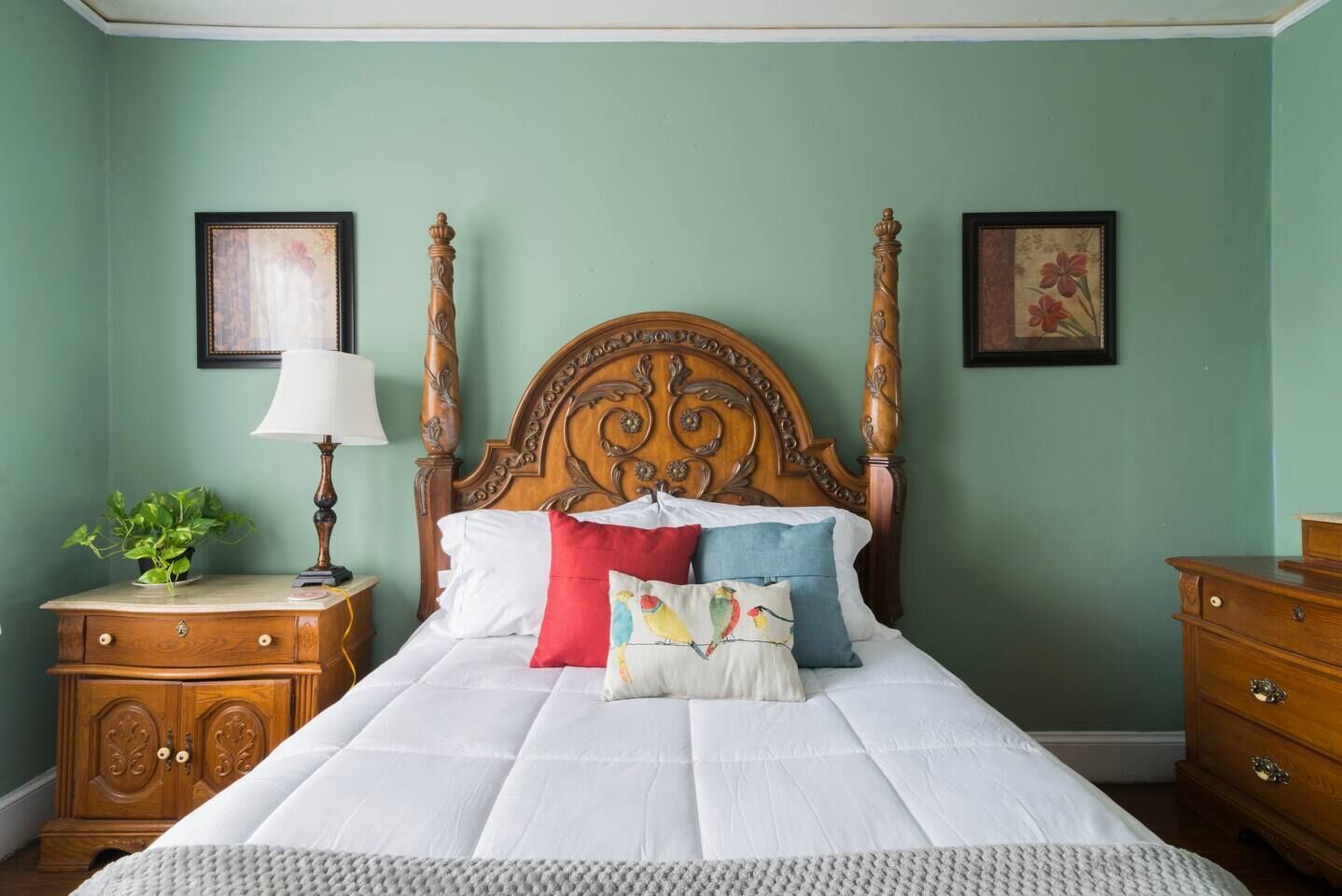 A bedroom with a four-poster bed, a nightstand with a lamp, a dresser, and two framed pictures on the wall.