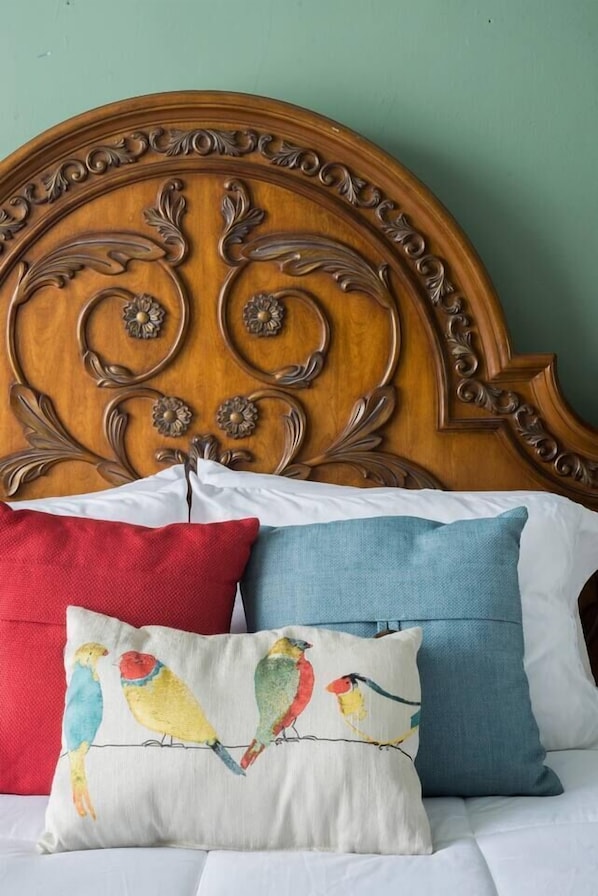 Un lit avec une tête de lit en bois ornée de motifs floraux sculptés et un coussin aux motifs colorés représentant des oiseaux.