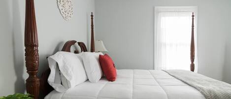 Una camera da letto con un letto a baldacchino, biancheria da letto bianca, un cuscino rosso, un comodino con una pianta e una finestra con tende leggere.