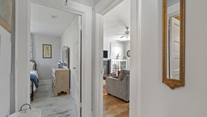 En gang der fører til et soveværelse med en seng, en kommode og et natbord. Der er en grå sofa i opholdsstuen med en loftsvifte.