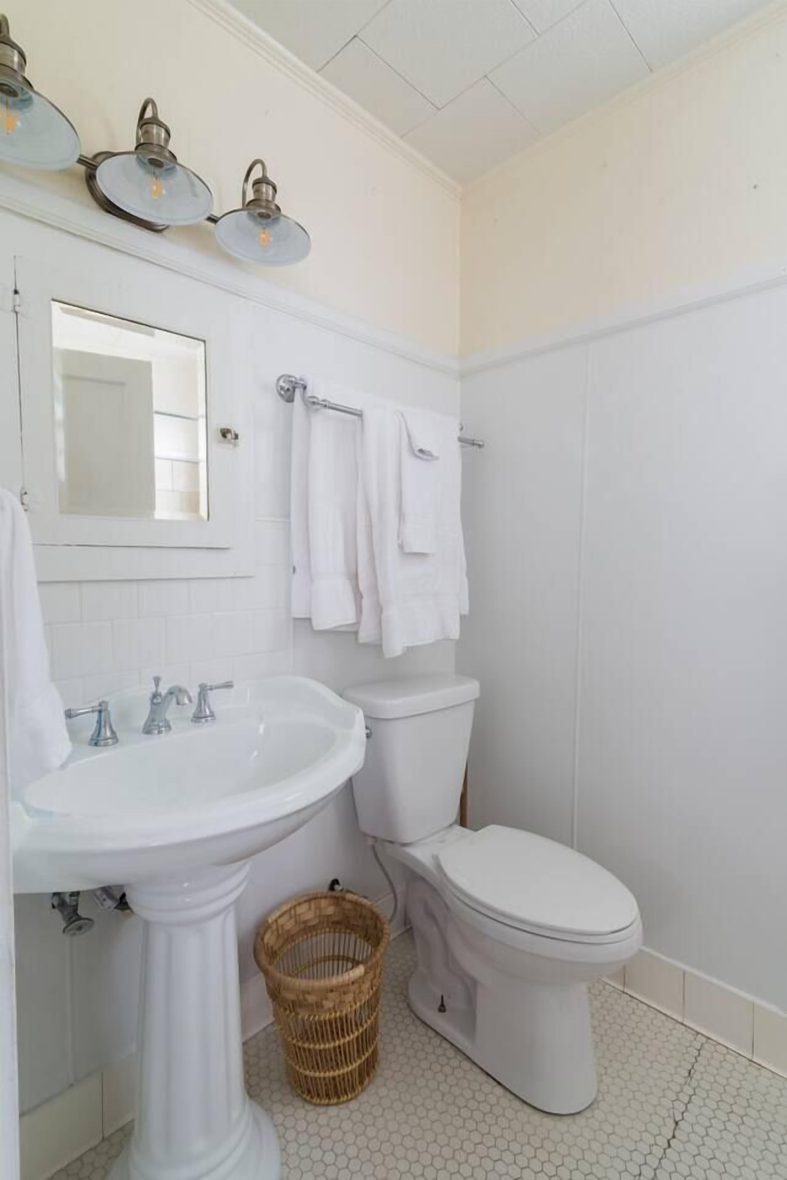 A bathroom with a pedestal sink, a toilet, and a mirror above the sink. There are three wall-mounted light fixtures above the sink.