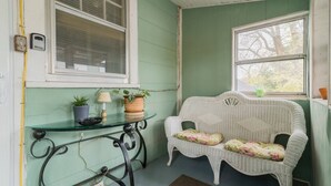 A room with a green wall, a white wicker sofa with floral cushions, a glass table with a lamp and potted plants, and a window with blinds.