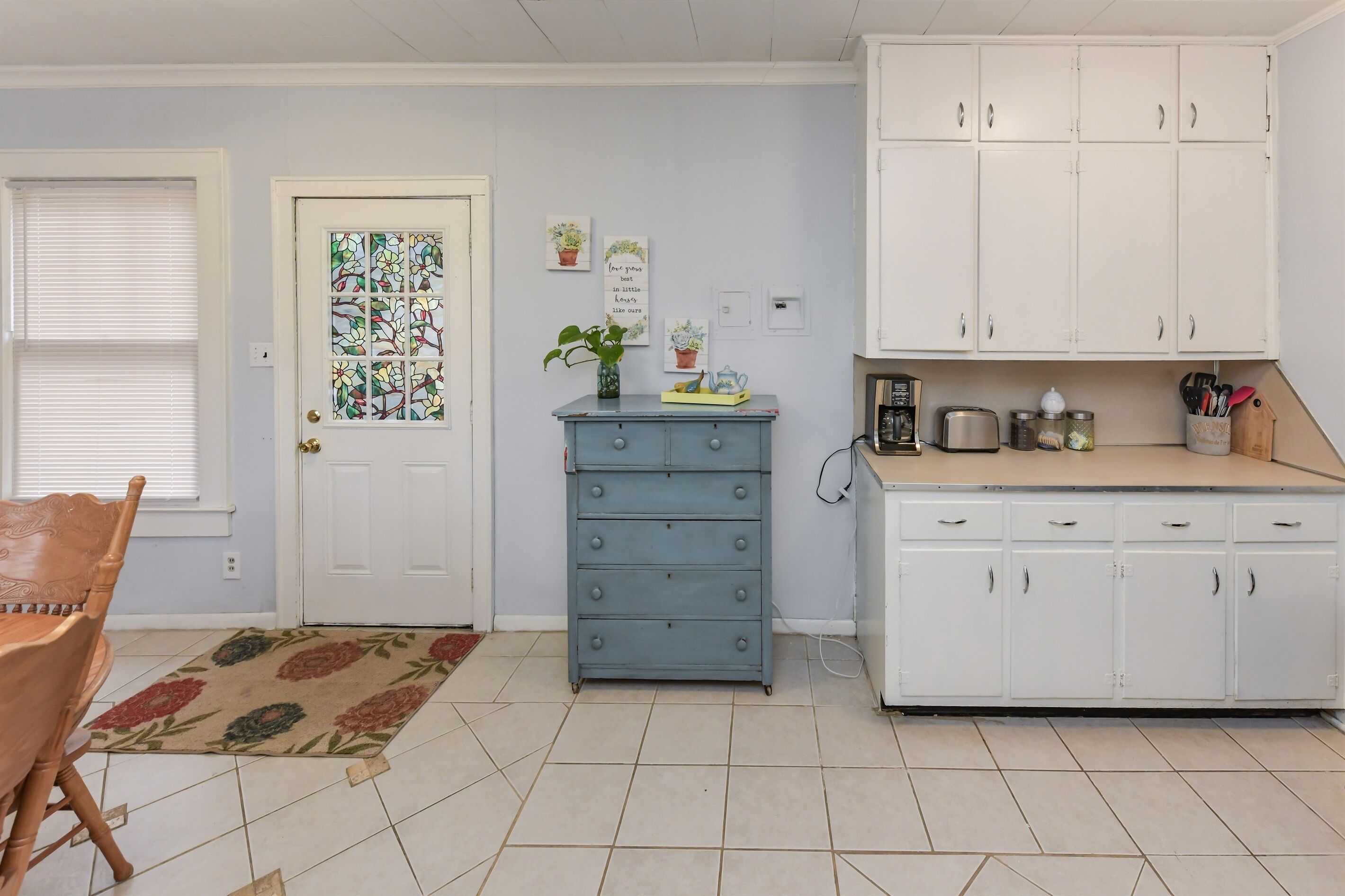 Une cuisine avec des meubles blancs, une commode bleue et une porte ornée d’un vitrail.