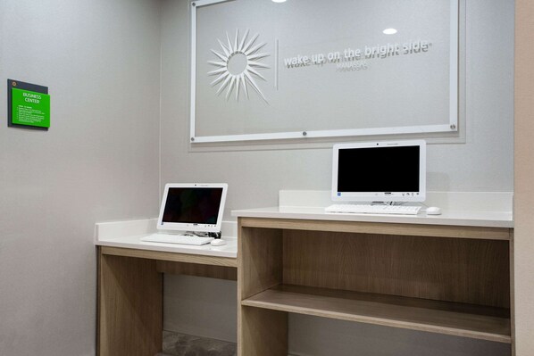 A business center with two white desks, one with a laptop and the other with a monitor, and a motivational poster on the wall.