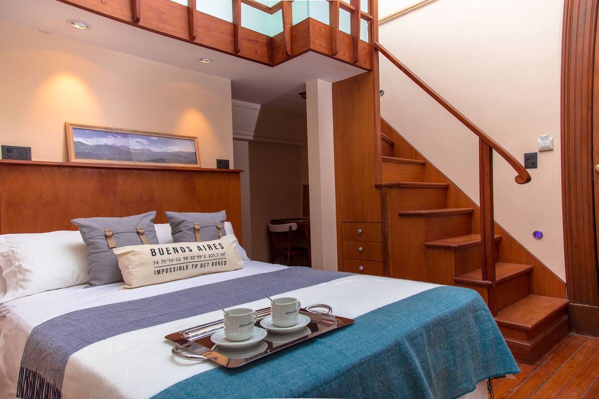 Quarto de hotel com cama, bandeja com xícaras e pires, escada de madeira e um quadro na parede.