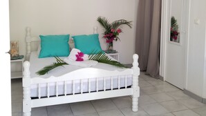 A white bed with teal pillows, a small bedside table with a vase of flowers, and a white ceiling with an air conditioning unit. - Studios Paradis Turquoise, swimming pool, beach, Les Trois ilets (les 3 ilets)