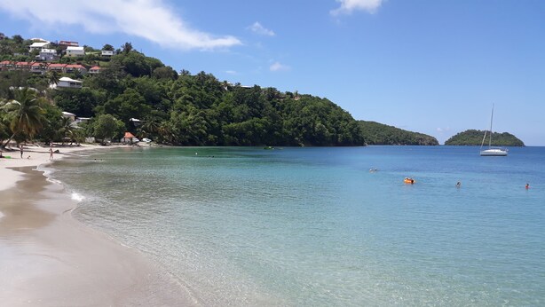 A beach with clear water, a sandy shore, and a hilly backdrop with buildings. - Studios Paradis Turquoise, swimming pool, beach, Les Trois ilets (les 3 ilets)
