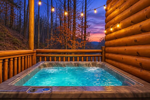 A hot tub with a wooden deck and string lights, surrounded by trees and a sunset.