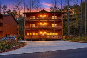 A multi-story log cabin with a balcony, surrounded by trees and a parking area.