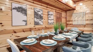 Une pièce aux murs lambrés, avec une longue table à manger dressée pour un repas, des œuvres d’art encadrées et une rangée de chaises.
