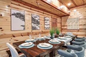 A wooden-paneled room with a long dining table set for a meal, framed artwork on the walls, and a row of chairs. - 6BR Cabin, All Ensuite | Indoor Pool + Theater (Pigeon Forge)