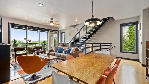 A spacious living area with a wooden dining table, modern chairs, a sofa with cushions, a staircase, and large windows offering a view of greenery. - The Abshier - Near U of A - Quiet Getaway (Fayetteville)