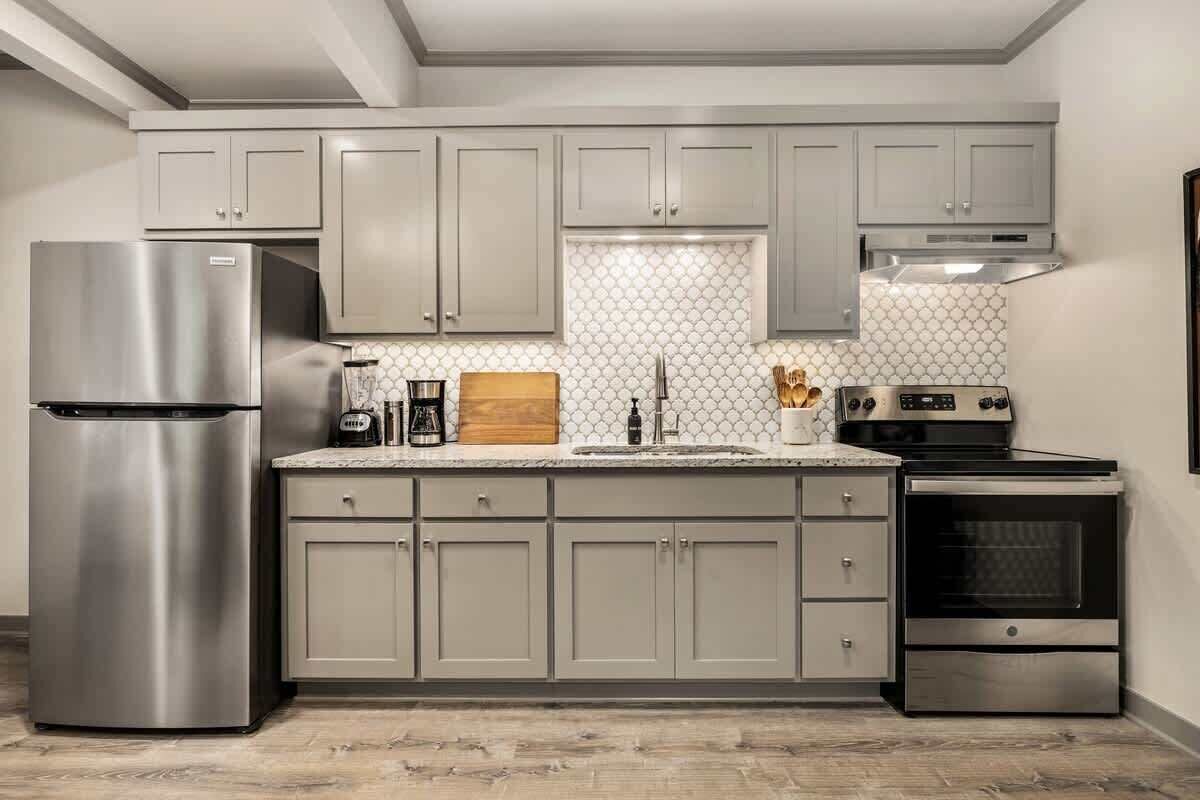 A modern kitchen with grey cabinets, a stainless steel refrigerator, a built-in oven, and a tiled backsplash.