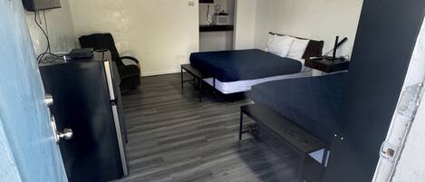 A compact room with a bed, a desk, a chair, and a small kitchenette.