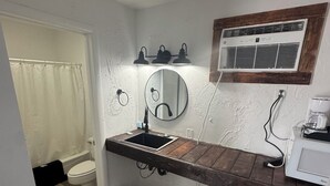 A bathroom with a wooden vanity, a round mirror, a sink, a toilet, and an air conditioning unit. - Holiday Lodge Motel (Sheridan)