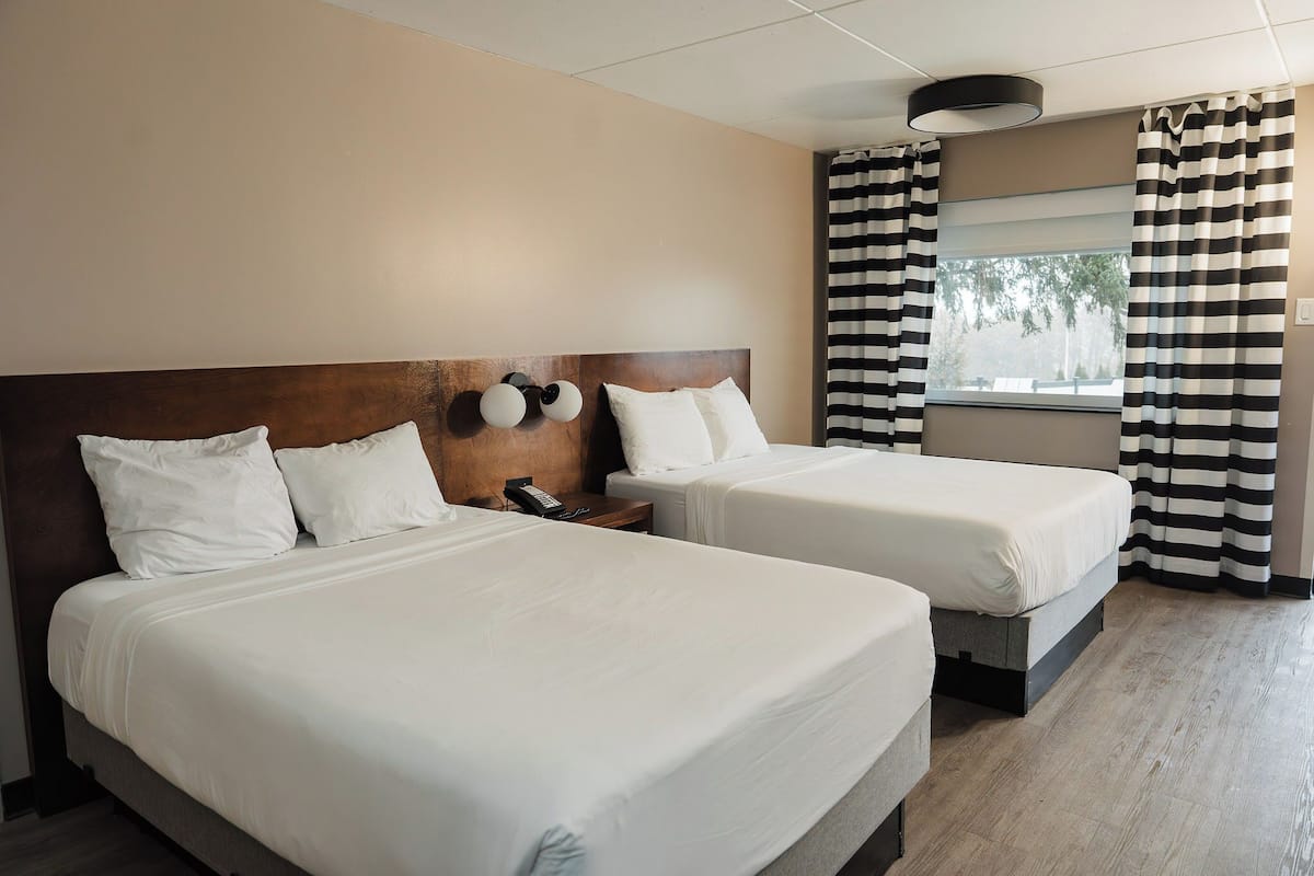 A hotel room with two beds, a wooden headboard, a bedside table with a phone, and a window with striped curtains.