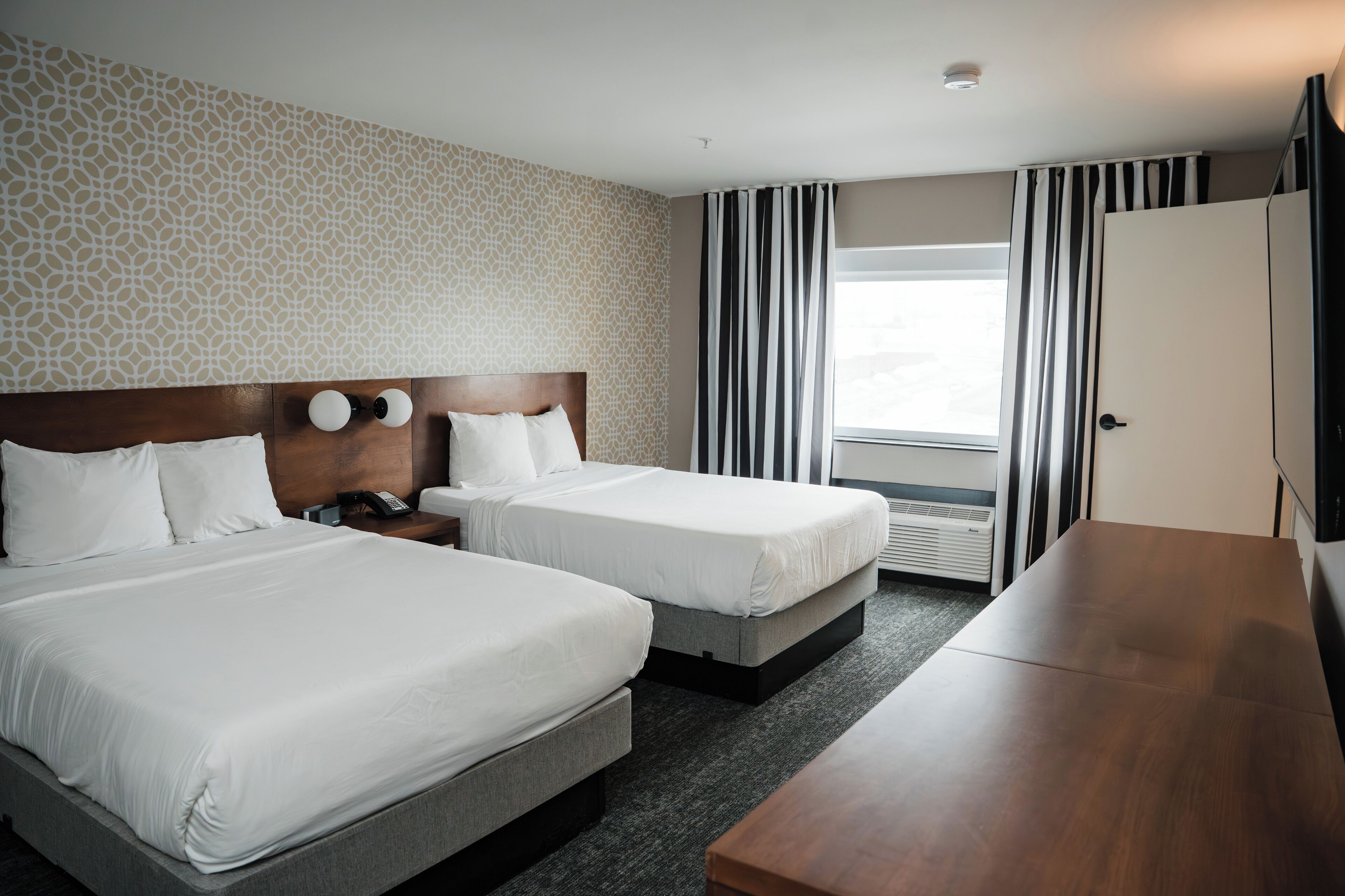 A hotel room with two beds, a wooden headboard, a desk, and a large window with curtains.