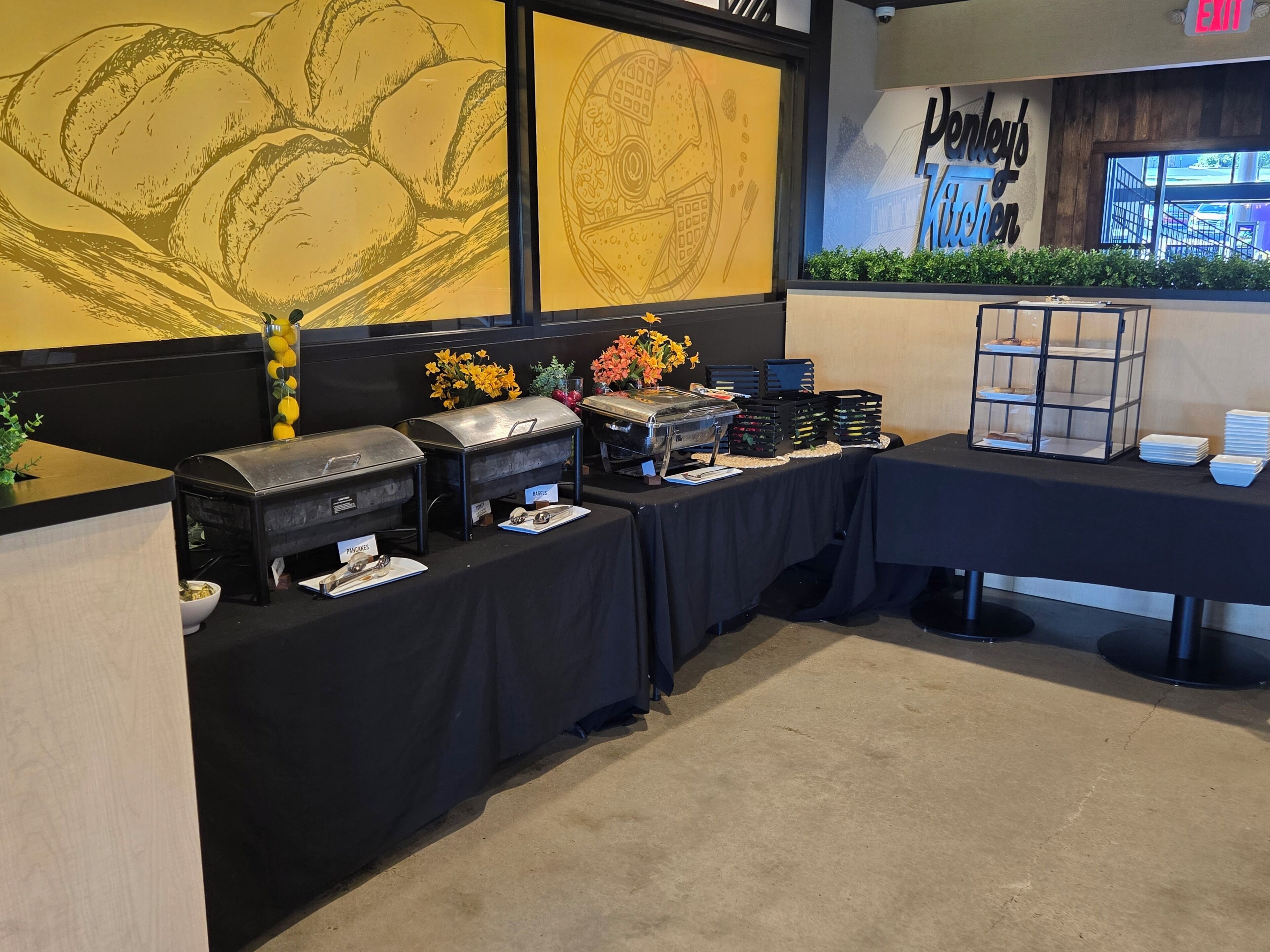 A buffet setup with labeled containers, a glass display case, and a wall with a bakery-themed mural.