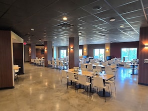 A spacious indoor dining area with multiple tables and chairs, large windows, and a grid-patterned ceiling.