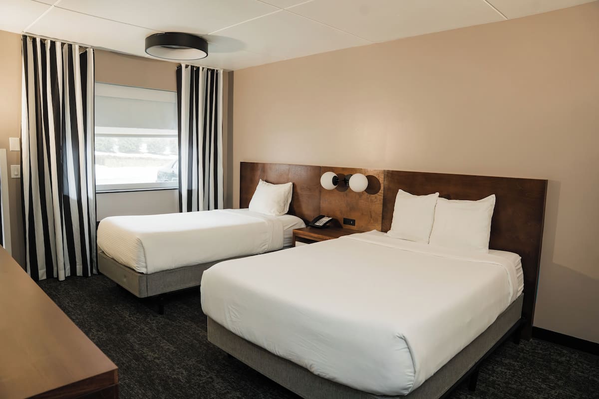 A hotel room with two beds, a wooden headboard, a bedside table with a phone, and a window with curtains.